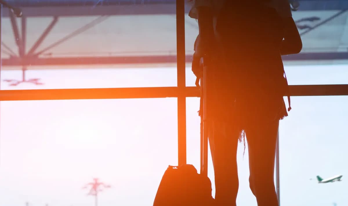 Silhuet af kvinde, der står med bagage i lufthavnsterminal ved solnedgang
